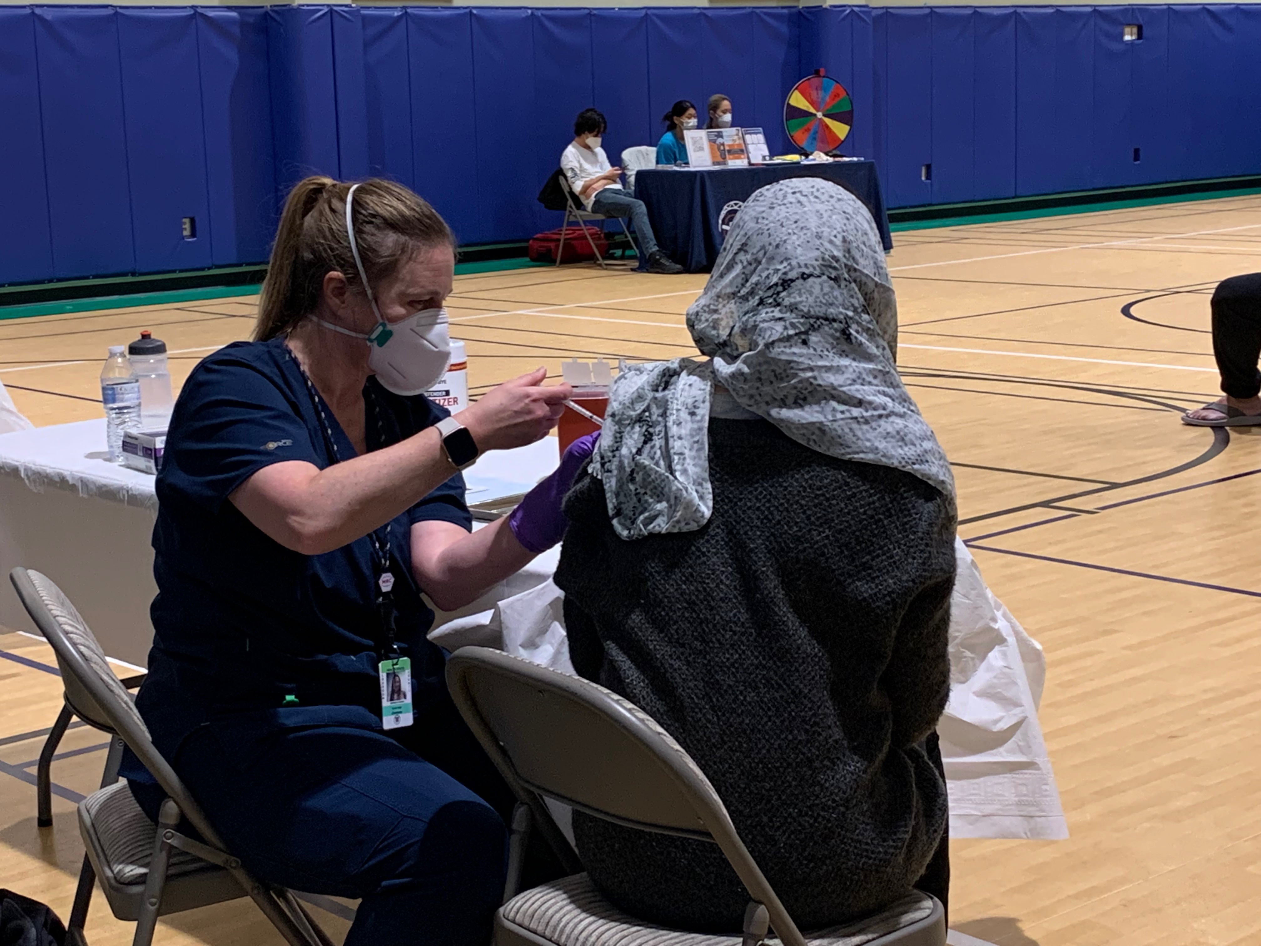 A healthcare worker administers a vaccination into the upper left arm of a person wearing a hijab at a table in an indoor gym.