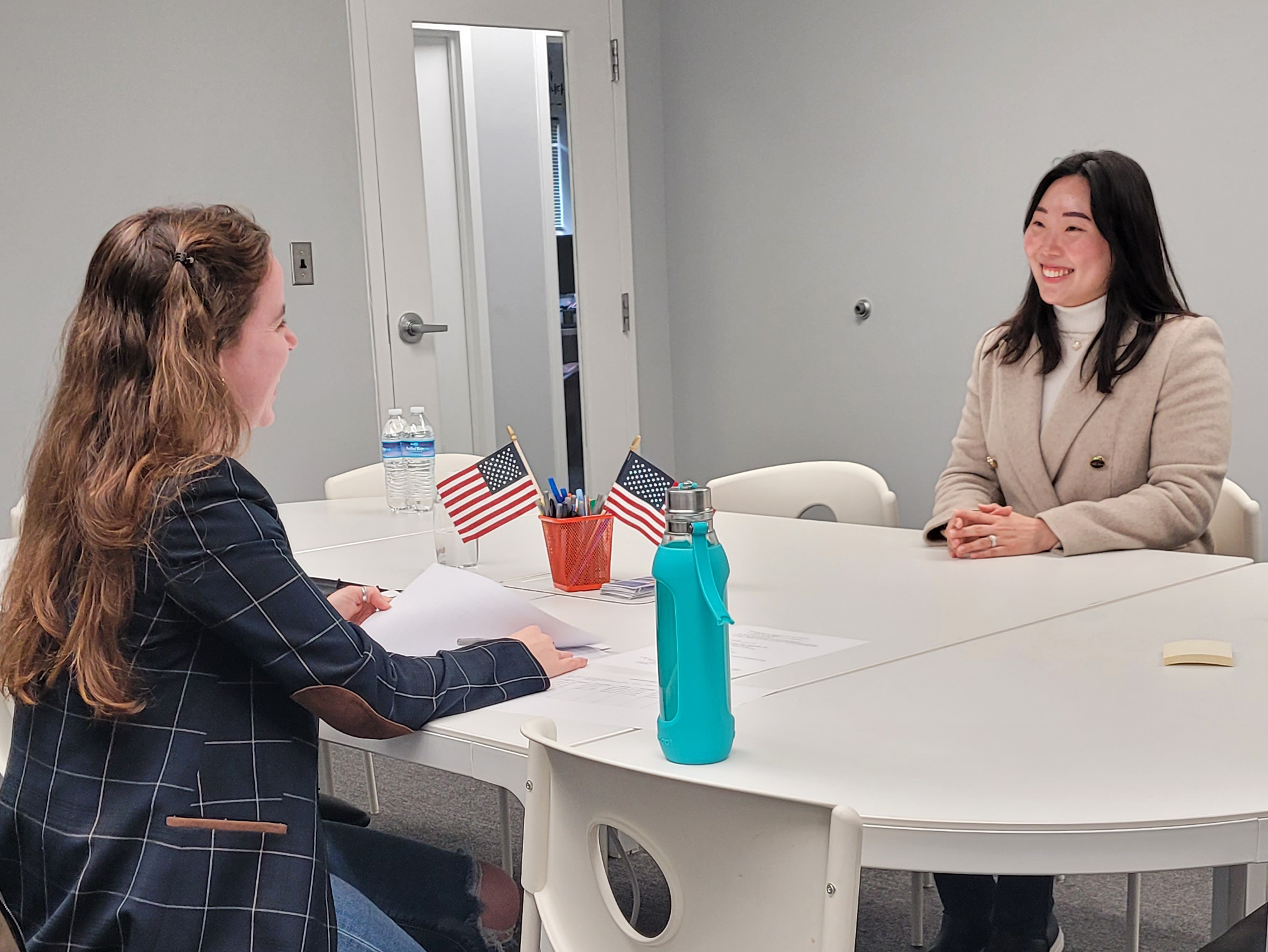 A person interviews another person at a white table, with 2 small US flags sticking out from a pencil holder.
