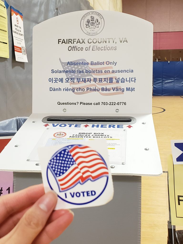 A person's hand holds up an "I VOTED" sticker, with the Fairfax County absentee ballot dropbox at a polling place in the background.