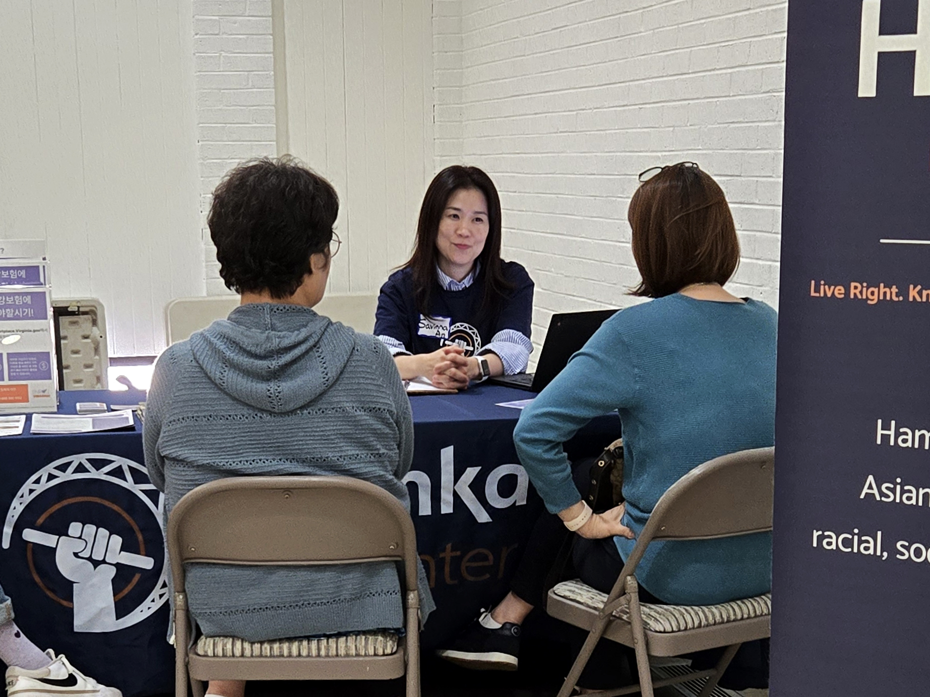 Savinna, Hamkae Center staff, speaks with 2 Korean community members at a table during a Hamkae Center services tabling event.